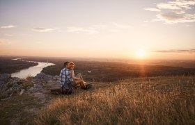 Ein Paar sitzt auf einem Hügel und blickt bei Sonnenuntergang auf einen Fluss und die Landschaft.