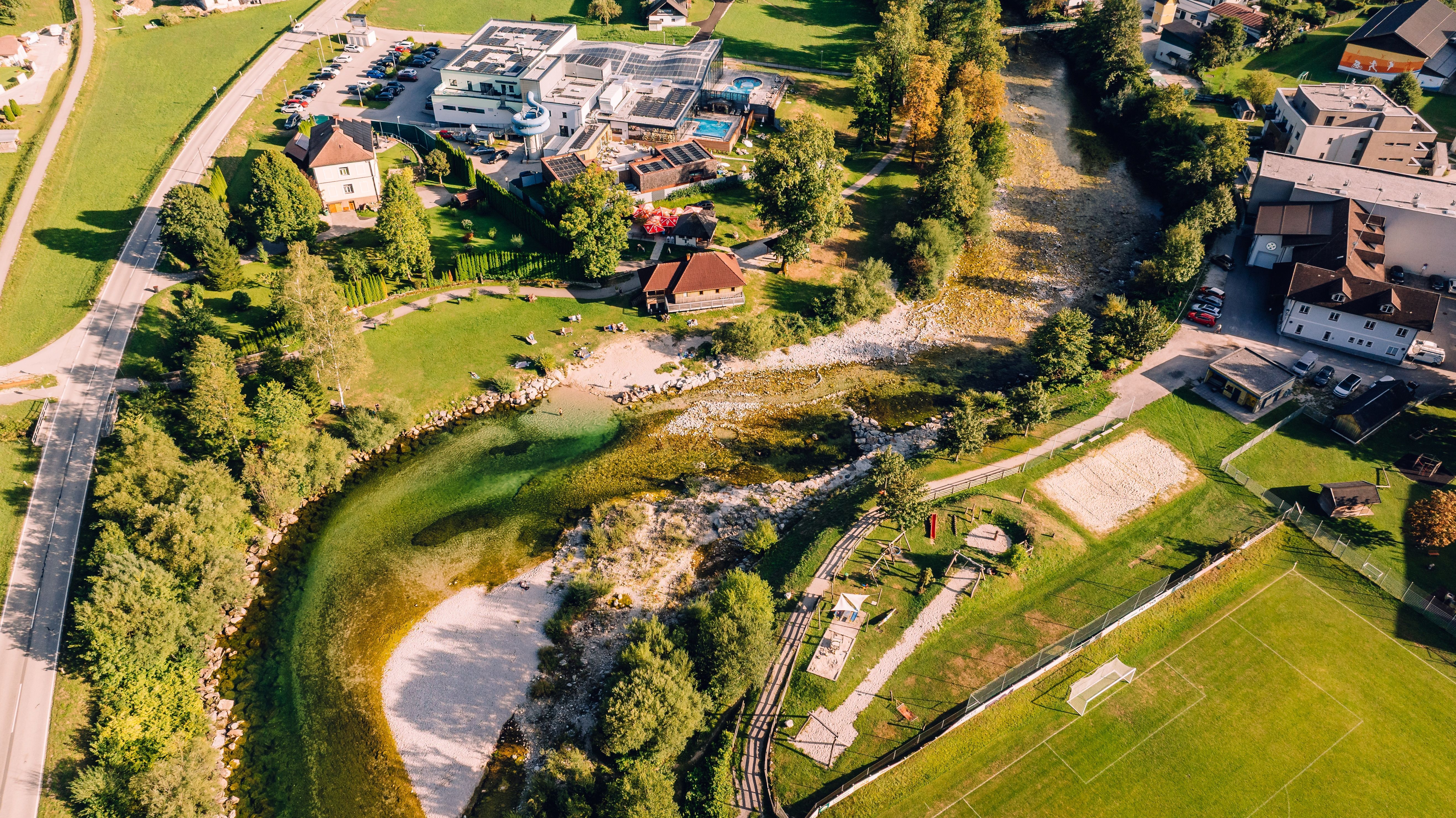 Luftaufnahme eines Flussufers mit Hotelanlage, Parkplätzen, Wiesen und Fußballplatz neben einem flachen, steinigen Flusslauf.