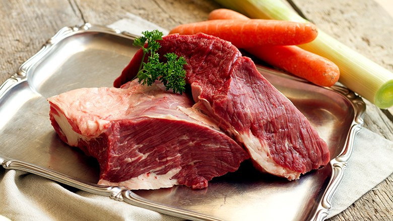 Zwei St&uuml;ck rohes Fleisch auf einem silbernen Tablett auf einem Holztisch. Daneben Kartotten und Lauch.