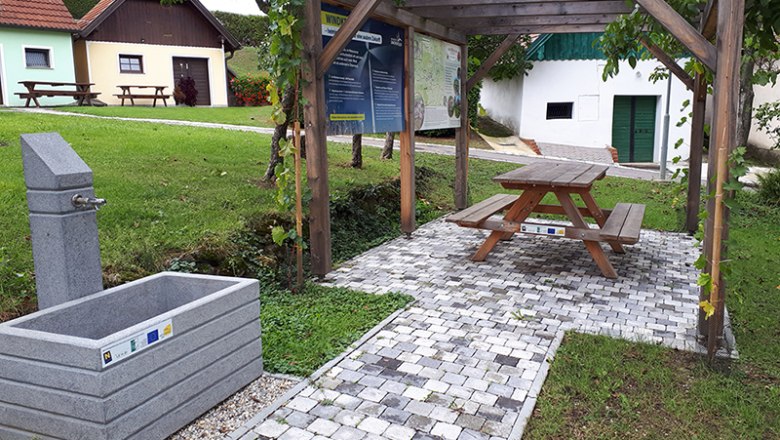 Ein Rastplatz mit Trinkbrunnen, Holztisch und Sitzbank unter einer Pergola, umgeben von gr&uuml;ner Wiese und kleinen Geb&auml;uden.