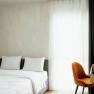 Modernes Hotelzimmer mit Doppelbett, weißen Bettlaken, Holzregal und orangefarbenem Stuhl vor einem Fenster mit Vorhängen.