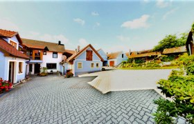 Hof mit mehreren Gebäuden, blauer Himmel, gepflasterter Boden, Blumen und Pflanzen.