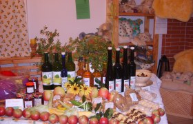 Ein Tisch in einem Hofladen mit verschiedenen Bio-Produkten wie &Auml;pfeln, Marmeladen, S&auml;ften und Lik&ouml;ren, dekoriert mit Blumen und Pflanzen.