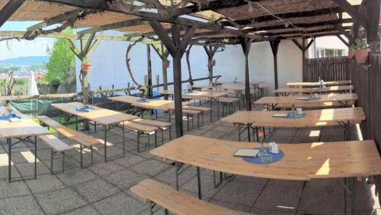 Leere Heurigenterrasse mit Holzb&auml;nken und Tischen unter einer Pergola.