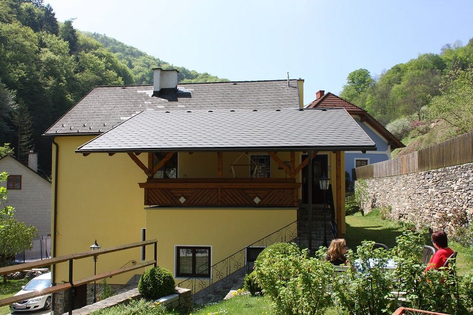 Gelbes Haus mit Balkon, umgeben von grüner Landschaft und Steinmauer.