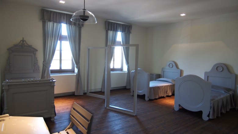 Ein schlicht eingerichtetes Schlafzimmer mit zwei Betten, einem Tisch und St&uuml;hlen, alles in grauen T&ouml;nen gehalten.