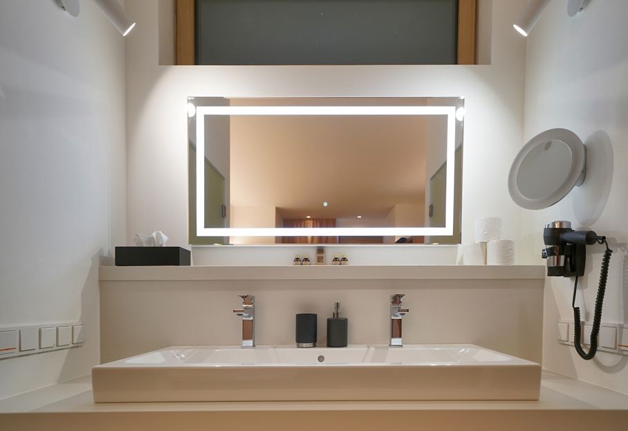 Moderner Waschtisch mit beleuchtetem Spiegel, zwei Wasserhähnen, Seifenspendern und Föhn.