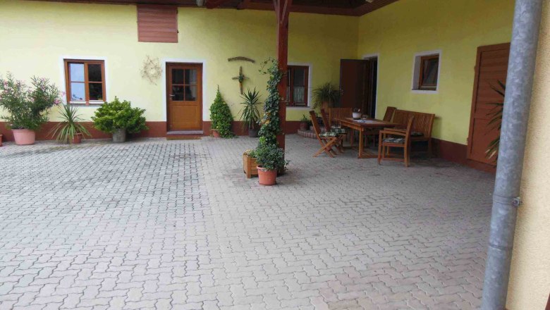 Innenhof mit gelber Fassade, Holztisch und St&uuml;hlen, umgeben von Pflanzen in T&ouml;pfen.