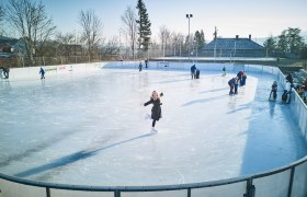Menschen beim Eislaufen auf einem Freiluft-Eislaufplatz mit Bäumen im Hintergrund.
