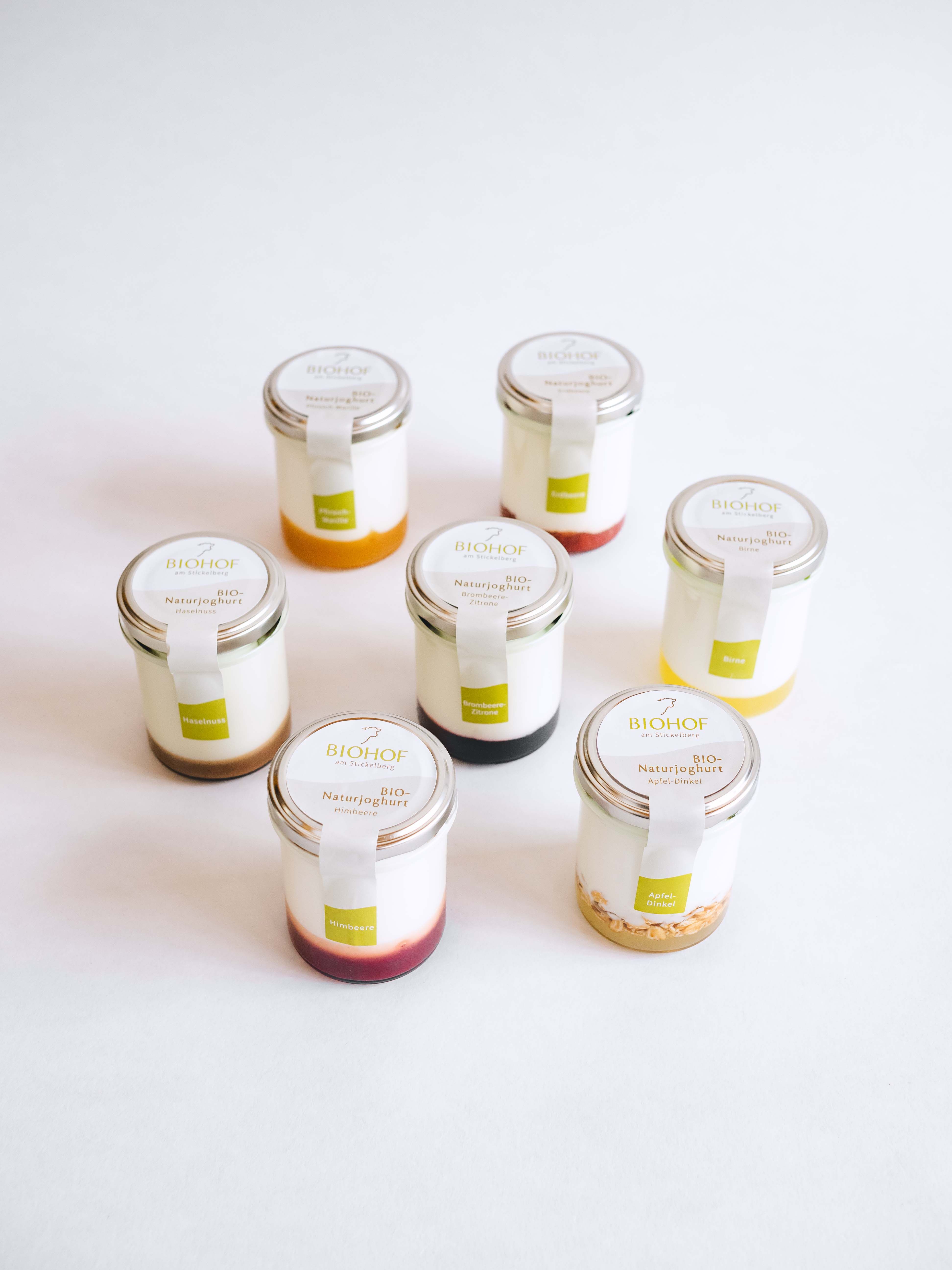 Sieben Gläser mit Naturjoghurt, jeweils mit unterschiedlichen Fruchtspiegeln, vor weißem Hintergrund.