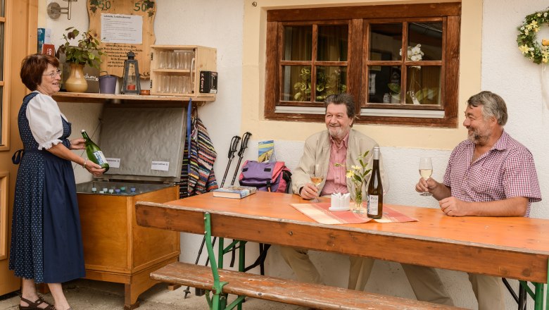 Drei Personen genießen Wein vor einem Haus, eine Frau in Tracht schenkt ein.