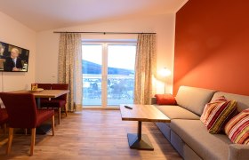 Gem&uuml;tliches Wohnzimmer mit roter Akzentwand, grauem Sofa, Holztisch, Fernseher und gro&szlig;em Fenster mit Vorh&auml;ngen.