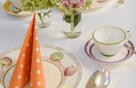 Ein elegant gedeckter Tisch mit Blumen, orange gepunkteten Servietten und Geschirr mit Obstmotiven.