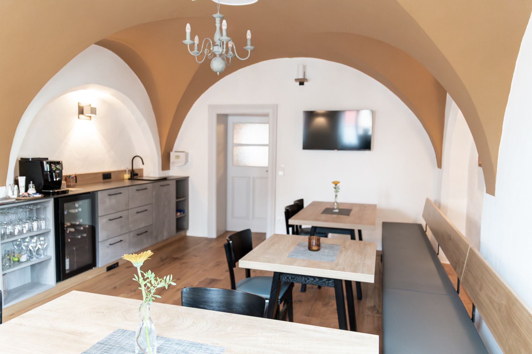 Ein gemütlicher Frühstücksraum mit Holztischen, Stühlen und einer Bank. An der Wand hängt ein Fernseher, und es gibt eine Küchenzeile mit Kaffeemaschine.