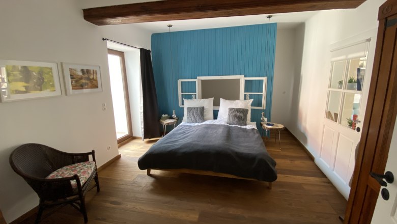 Modernes Schlafzimmer mit Holzboden, blauem Akzent an der Wand, Doppelbett und Rattan-Stuhl.