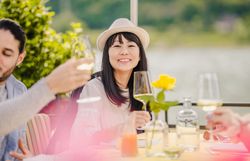 Eine Dame mit Hut sitzend an einem Tisch hält ein gefülltes Weißweinglas hoch.