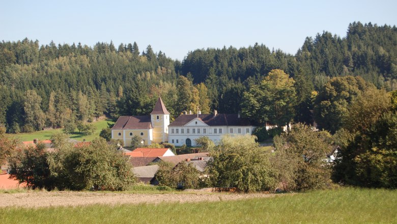 Ein Schloss mit gelbem Turm inmitten einer gr&uuml;nen Landschaft und umgeben von B&auml;umen.