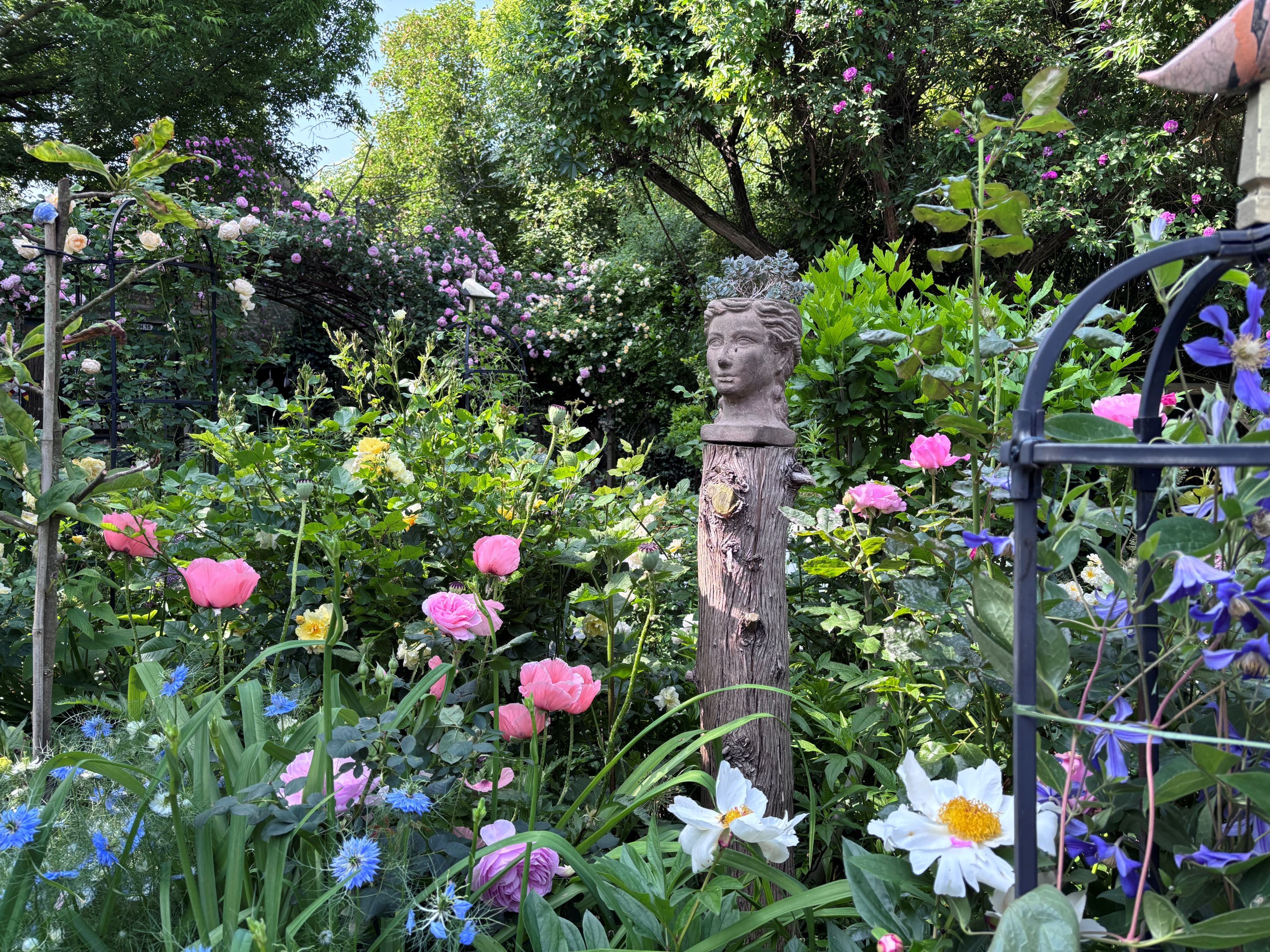 Ein blühender Garten mit bunten Blumen und einer Büste auf einem Holzpfosten.