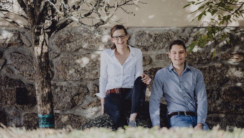 Zwei Personen sitzen l&auml;chelnd auf einem Stein vor einer Steinmauer, umgeben von B&auml;umen.