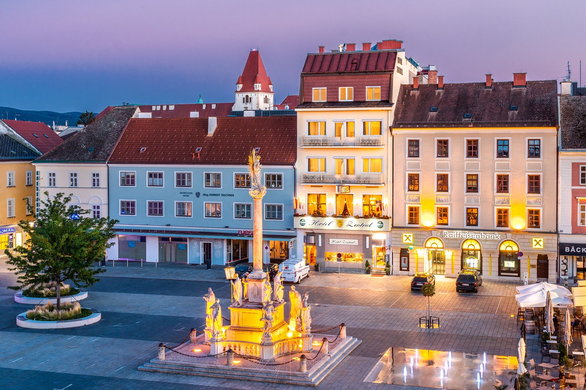 Abendliche Stadtansicht mit beleuchtetem Brunnen und Gebäuden, darunter ein Hotel und eine Bank.