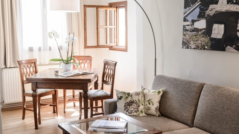 Gemütliches Wohnzimmer mit grauem Sofa, Holztisch und Stühlen, hellem Fenster und Pflanzen.