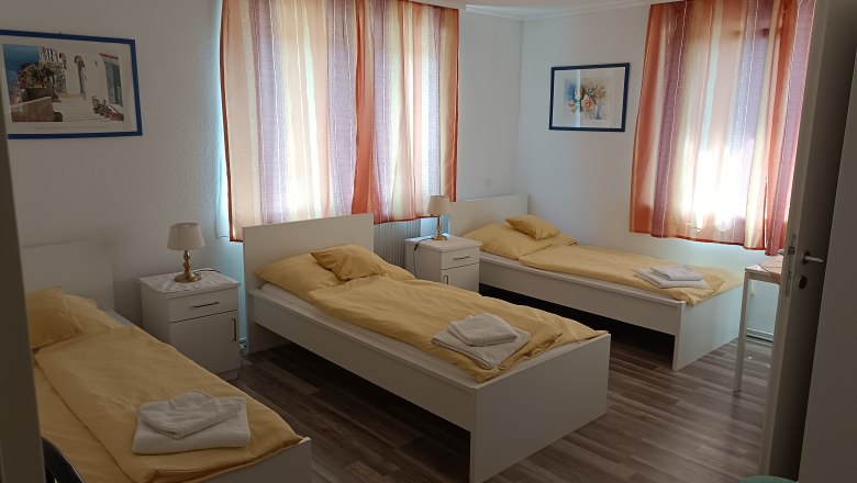 Drei Einzelbetten in einem hellen Zimmer mit gelber Bettw&auml;sche und bunten Vorh&auml;ngen.