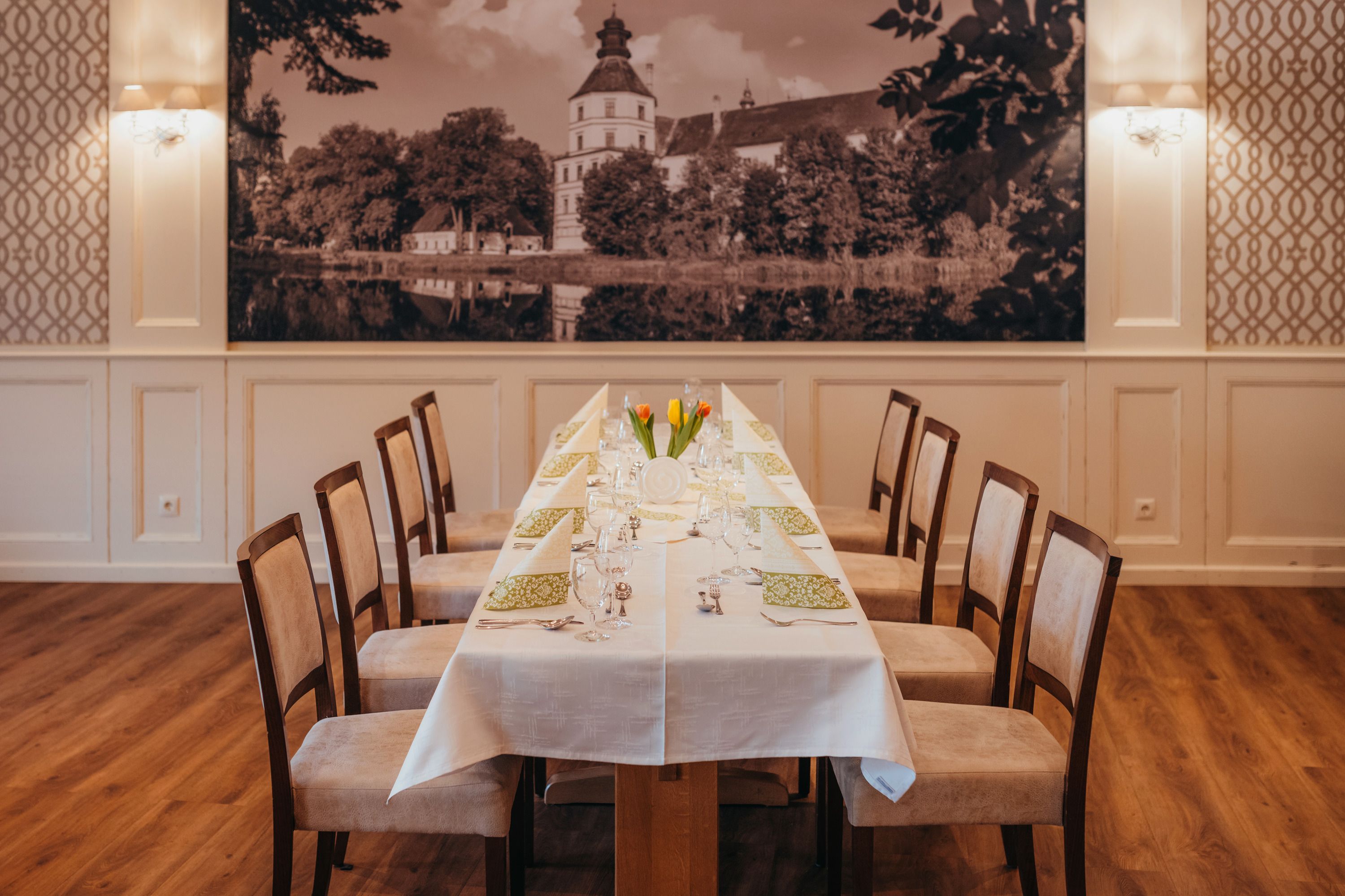 Eleganter Speisesaal mit langem Tisch, gedeckt mit weißen Tischdecken und grünen Servietten, im Hintergrund ein großes Schlossbild.