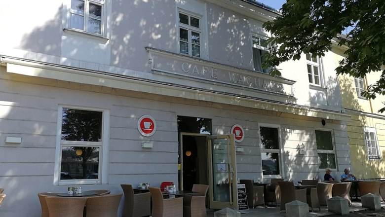 Au&szlig;enansicht eines Caf&eacute;s mit Terrasse und Sitzgelegenheiten unter einem Baum.