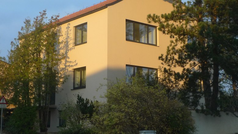 Ein modernes, dreist&ouml;ckiges Haus mit gelber Fassade und roten Dachziegeln, umgeben von B&auml;umen und Str&auml;uchern.