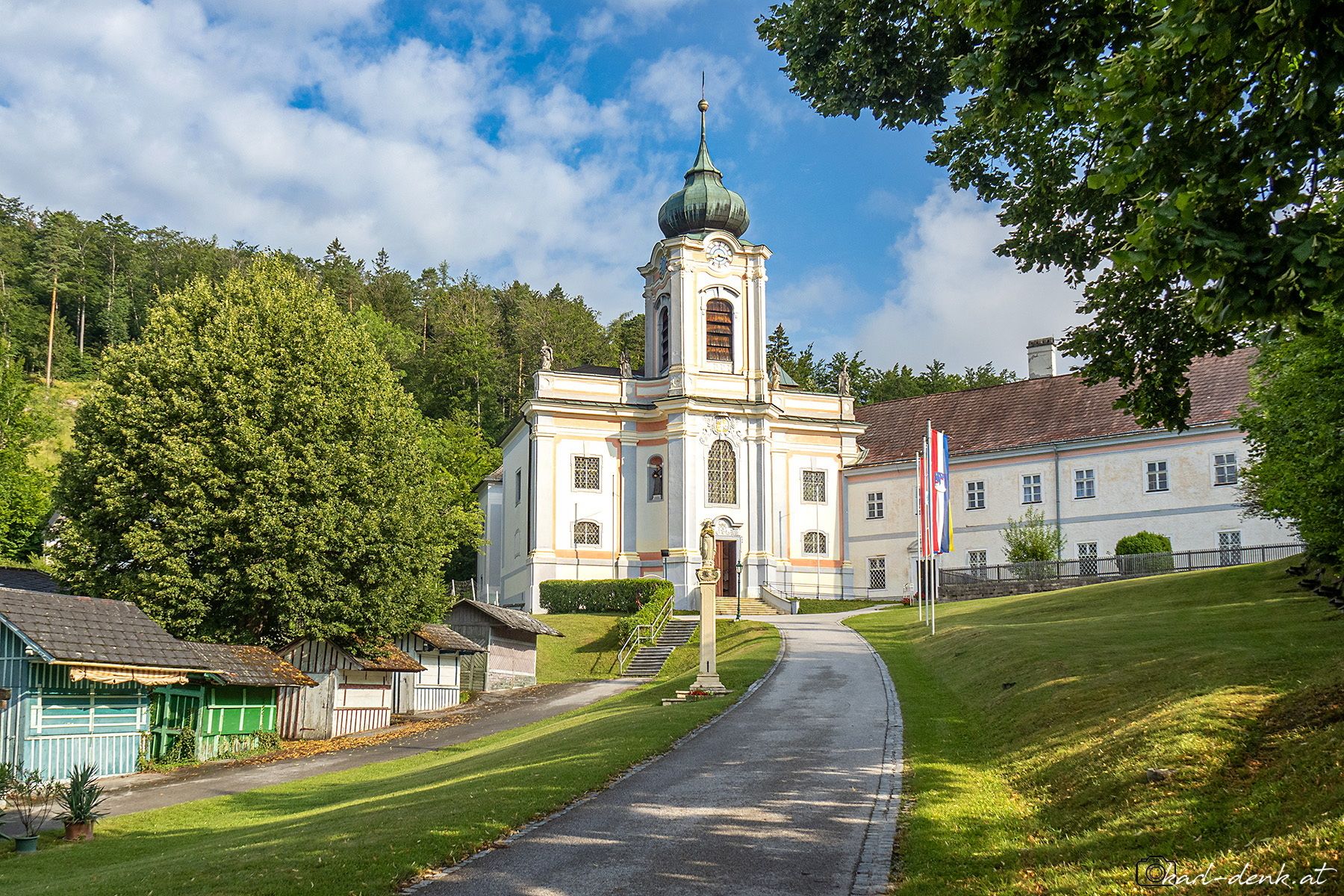 Das Servitenkloster Mariahilfberg in Gutenstein mit einer barocken Kirche und einem Turm, umgeben von Bäumen und einem Weg im Vordergrund.