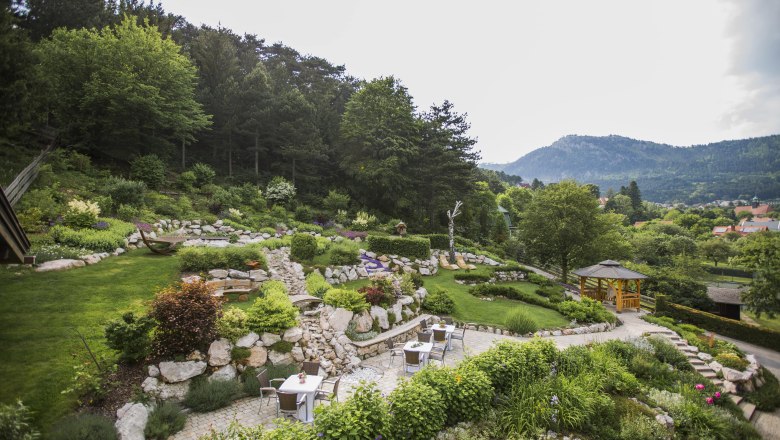 Ein terrassierter Garten mit Steinen, Pflanzen und Sitzbereichen, umgeben von Bäumen und Bergen im Hintergrund.