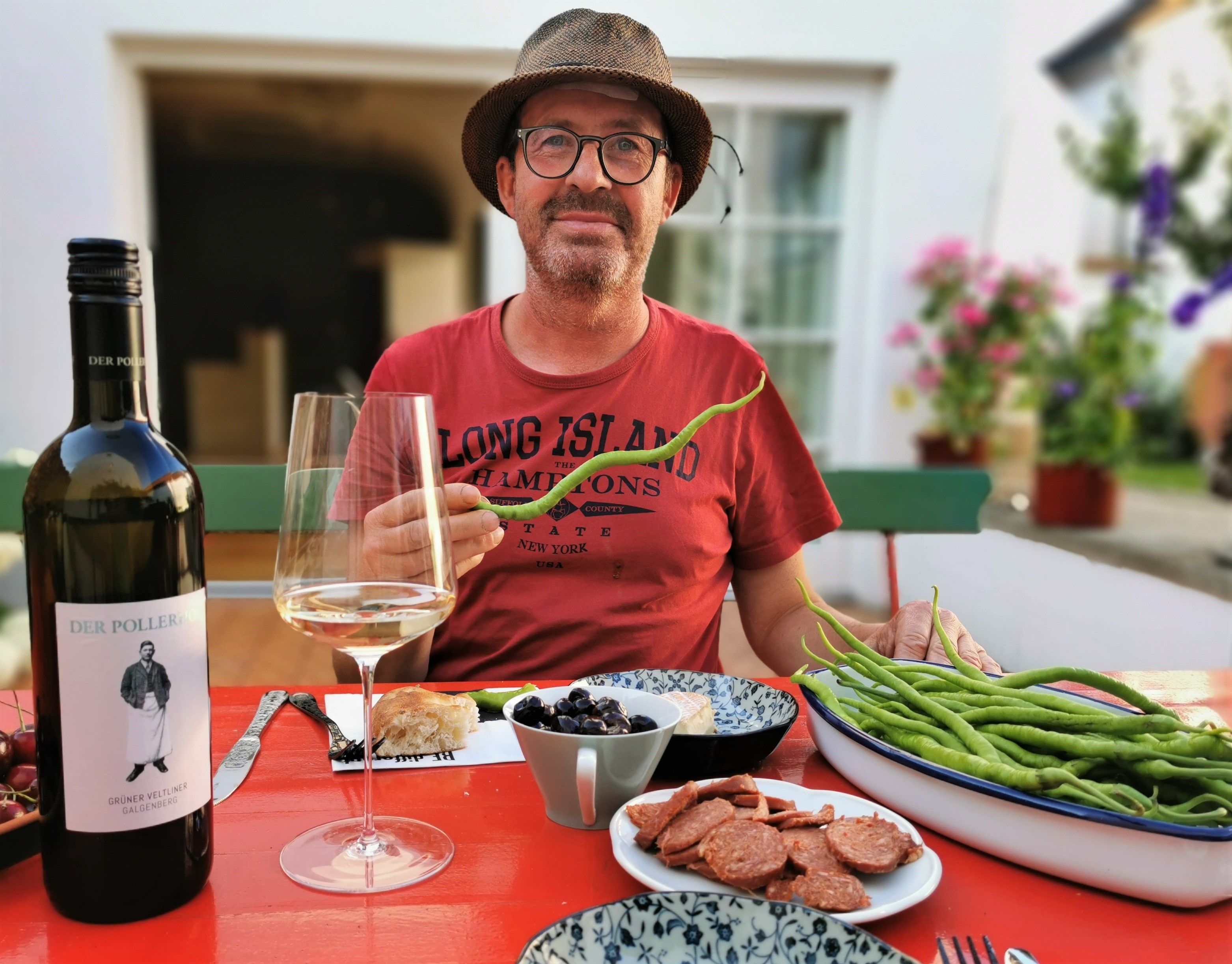 Ein Mann mit Hut sitzt an einem Tisch mit Weinflasche, Glas, Bohnen und Wurst.