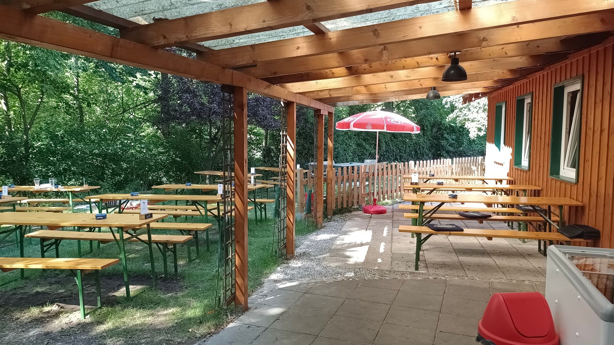 Überdachte Terrasse mit Holzbänken und Tischen, umgeben von Bäumen und einem Holzzaun.