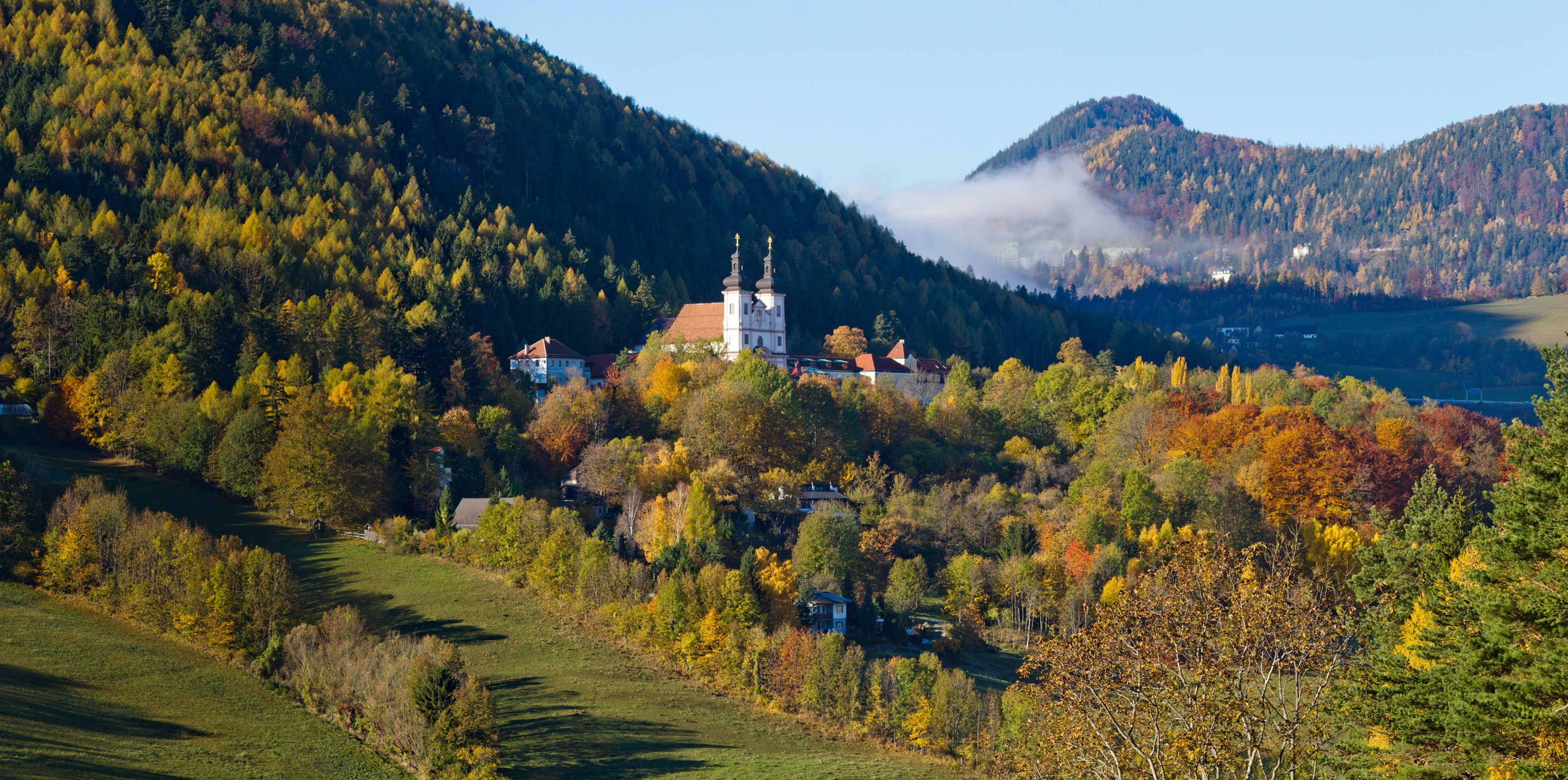 Herbstliche Landschaft mit Kirche in Maria Schutz, umgeben von bunten Bäumen und Hügeln.