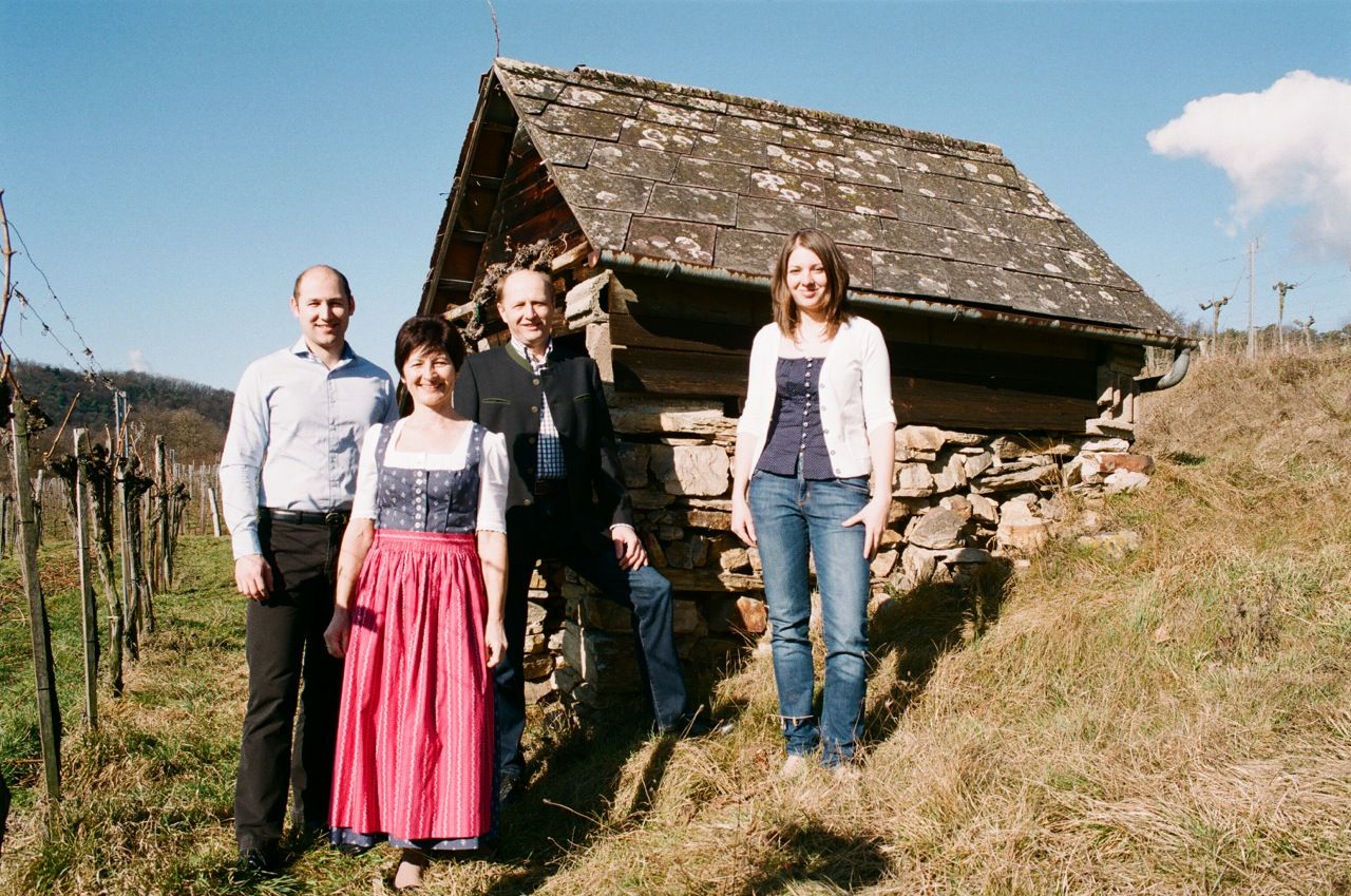 Vier Personen stehen vor einer alten Hütte auf einem Hügel mit Weinreben im Hintergrund.