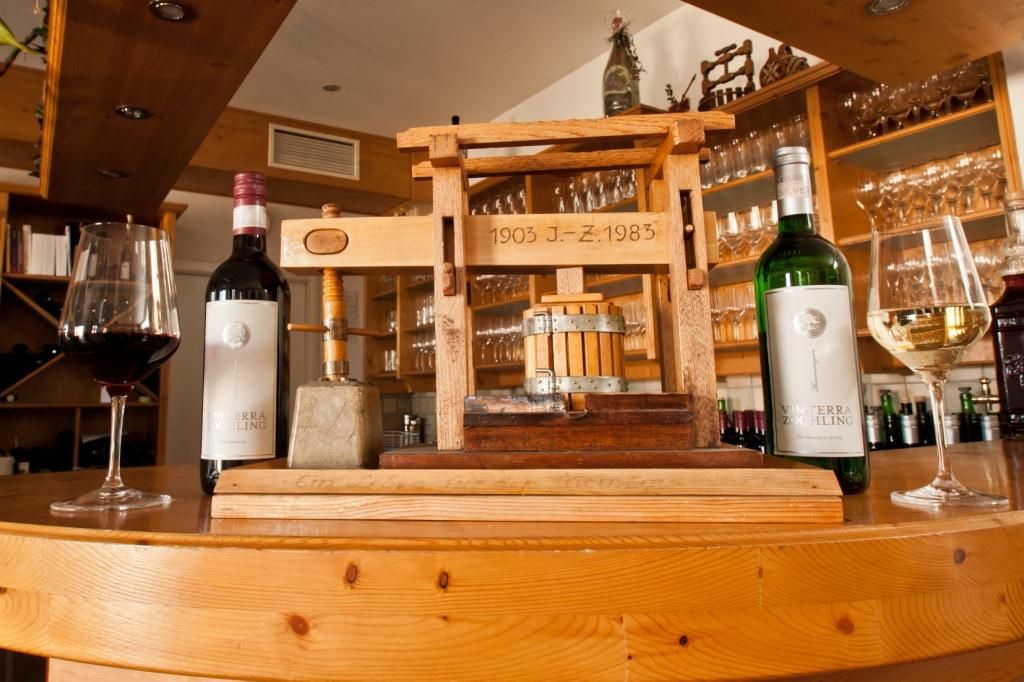 Weinflaschen und Gläser neben einer kleinen Holzpresse auf einer Theke.