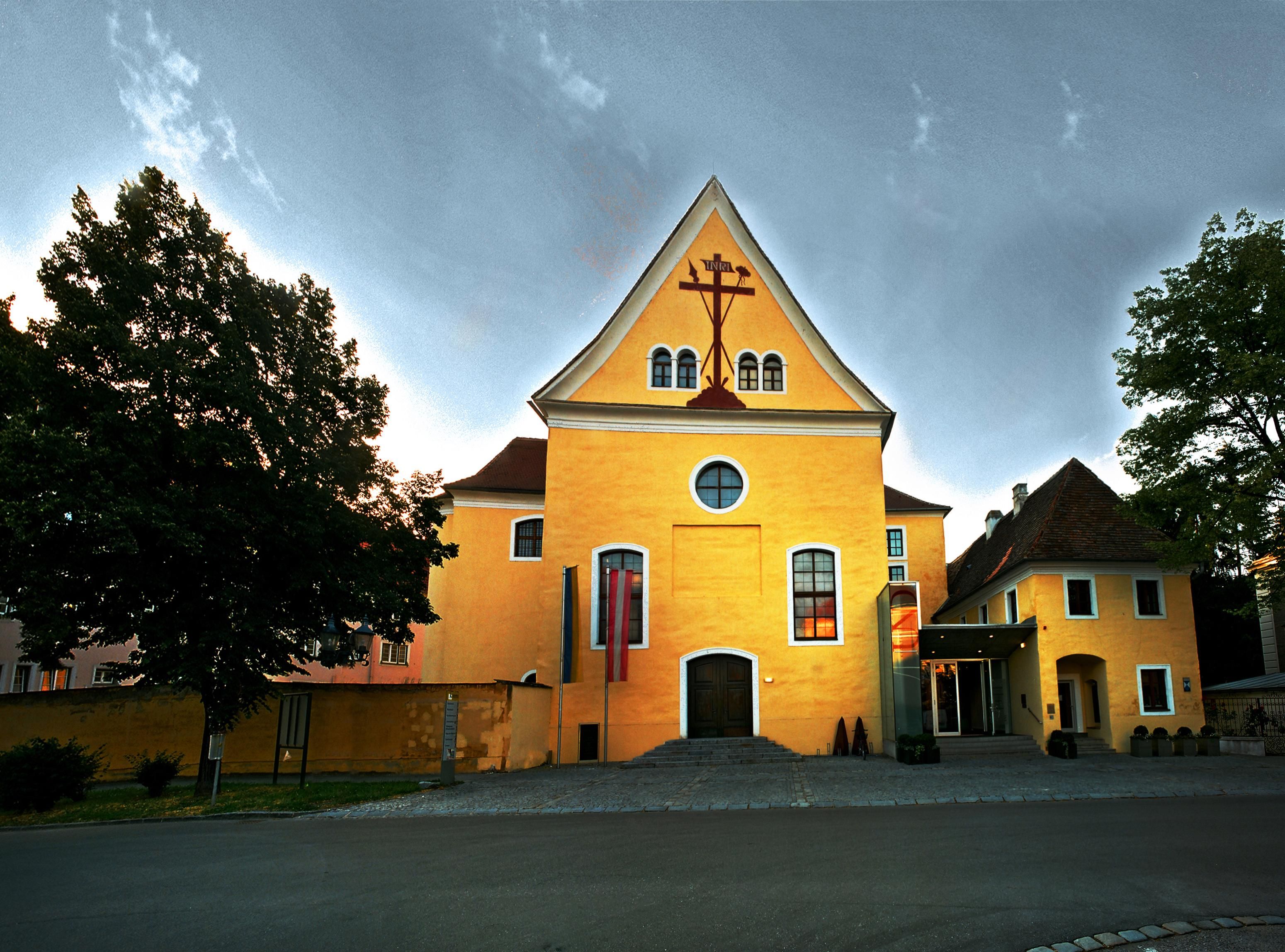 Gelbes Gebäude mit Kreuzsymbol und Fahnen vor dem Eingang.