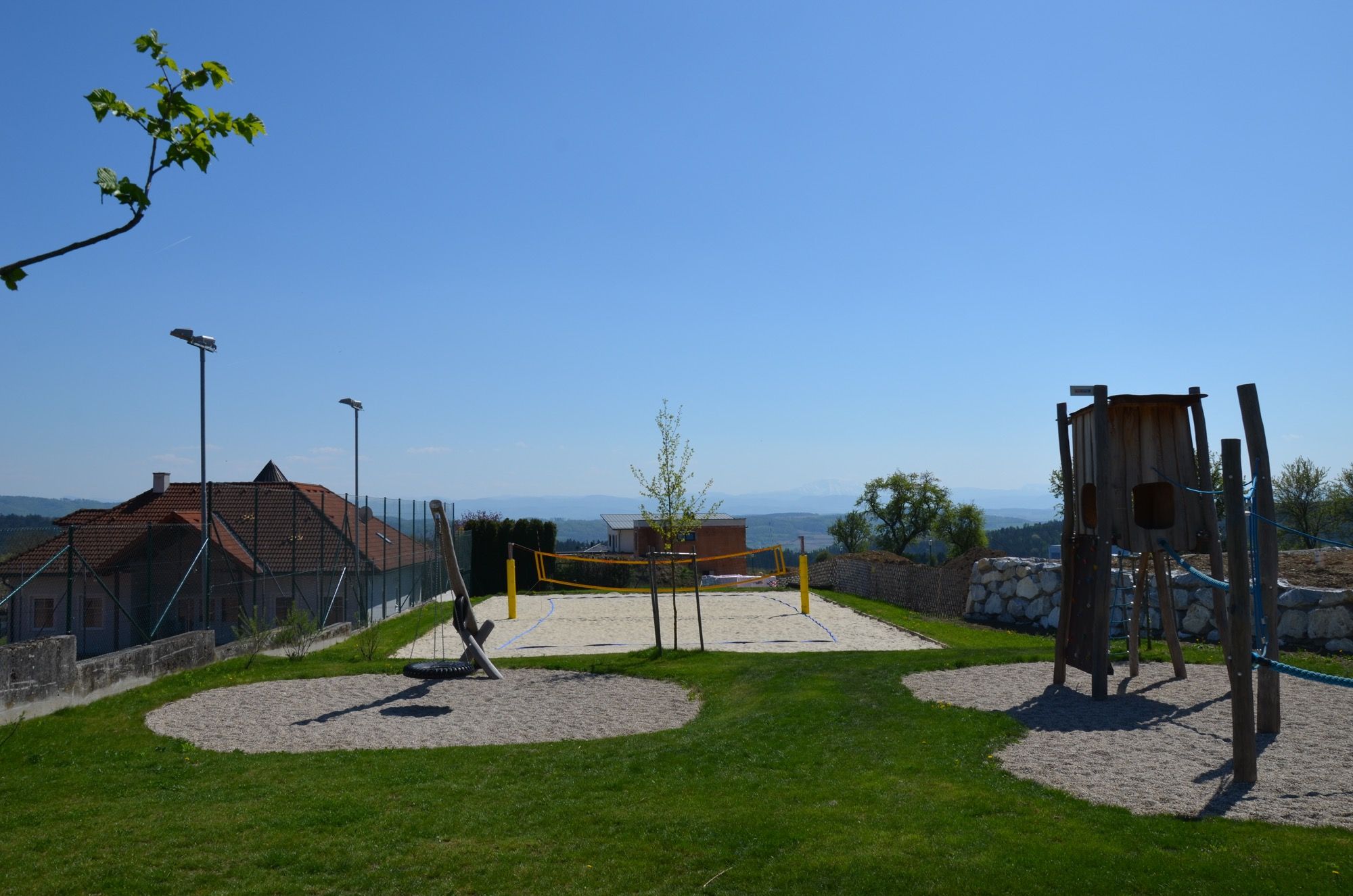 Ein Beachvolleyballplatz mit Netz, umgeben von grünem Rasen und einem Spielplatz im Vordergrund, unter klarem blauem Himmel.