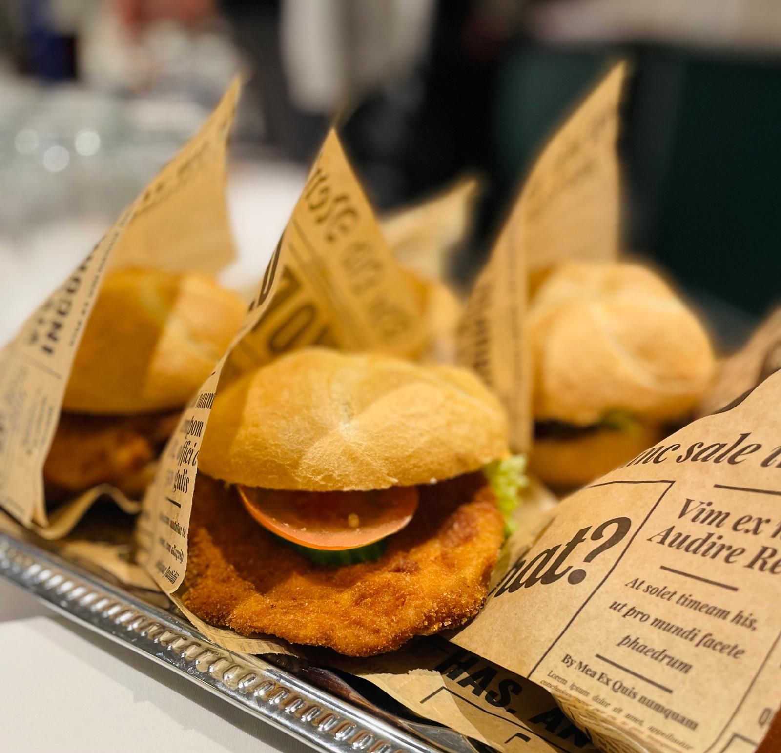 Mehrere Burger in Zeitungspapier auf einem Tablett, im Vordergrund ein Burger mit paniertem Patty, Tomate und Gurke.
