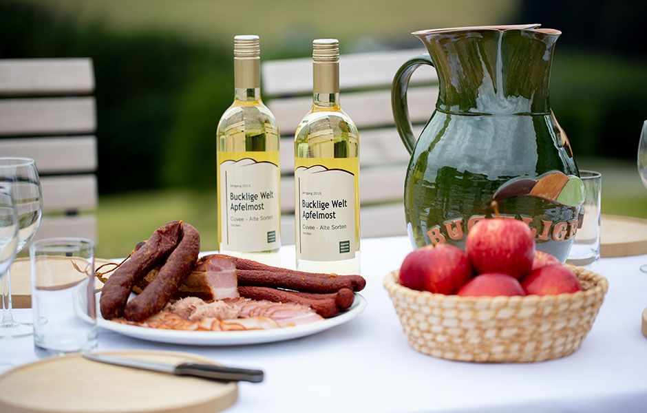 Tisch mit Apfelmost, Wurstplatte, Äpfeln, Holzbrett, Messer und Gläsern.