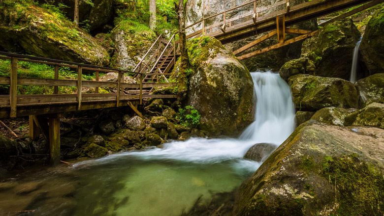 Holzbrücken und Wasserfall in einer bewaldeten Schlucht.