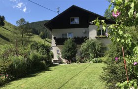 Ein zweistöckiges Haus mit Balkon, umgeben von einem gepflegten Garten und grünen Hügeln im Hintergrund.