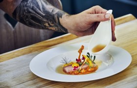 Ein Teller mit kunstvoll angerichtetem Essen, über das eine Hand aus einer kleinen Kanne Sauce gießt.