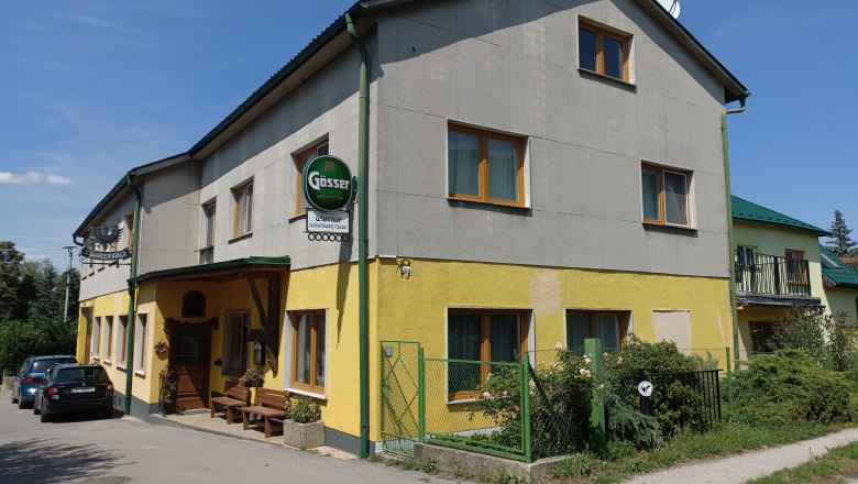 Au&szlig;enansicht eines gelb-grauen Gasthofs mit G&ouml;sserschild und parkenden Autos.