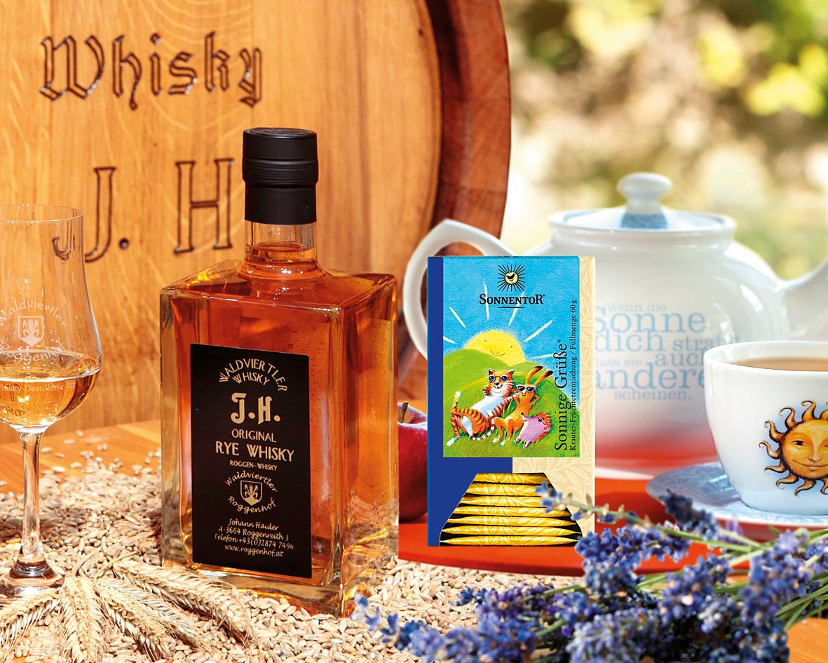 Eine Flasche Waldviertler Rye Whisky neben einem Glas, Teepackung und Teekanne auf einem Tisch mit Lavendel und Getreide.