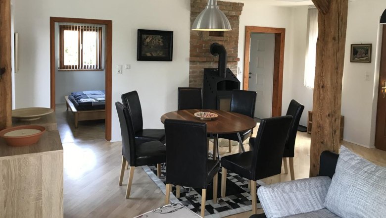 Essbereich mit rundem Holztisch, schwarzen St&uuml;hlen und Holzofen in einem Raum mit Holzbalken.