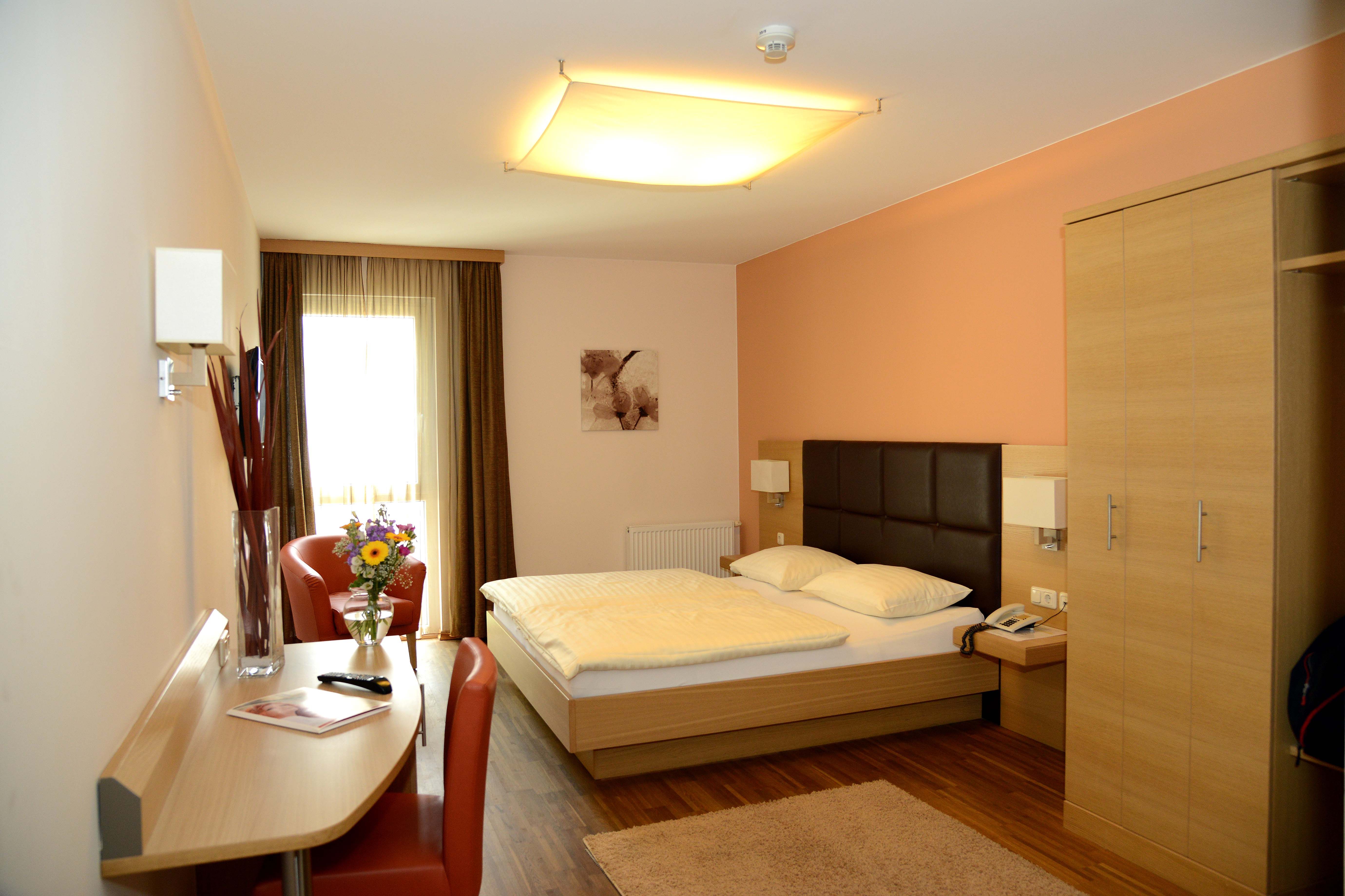 Ein modernes Hotelzimmer mit Doppelbett, Schreibtisch, Stuhl und Blumenstrauß auf einem Tisch.