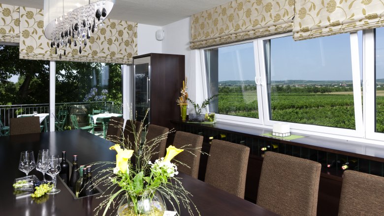 Ein Verkostungsraum mit einem gro&szlig;en Tisch, Weingl&auml;sern und Flaschen. Gro&szlig;e Fenster bieten Blick auf Weinberge. Elegante Dekoration und Beleuchtung.