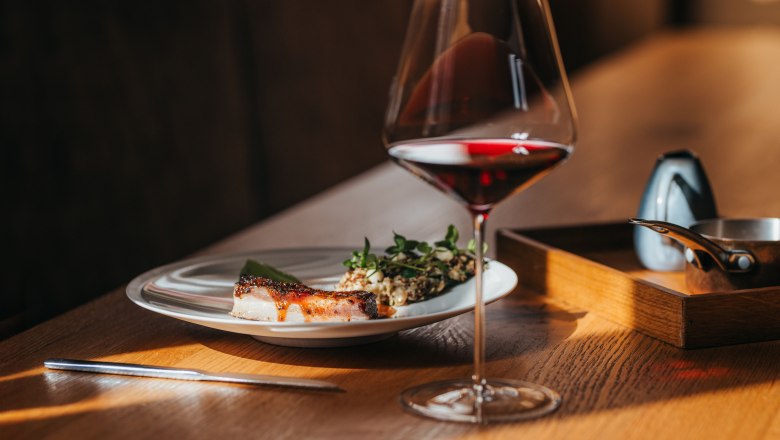 Ein Teller mit Lammbauch, Kopfsalat und Wasabicrème neben einem Glas Rotwein auf einem Holztisch.