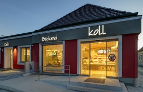 Außenansicht am Abend der Bäckerei mit beleuchtetem Schild 'Bäckerei Koll' und 'Cafe'.